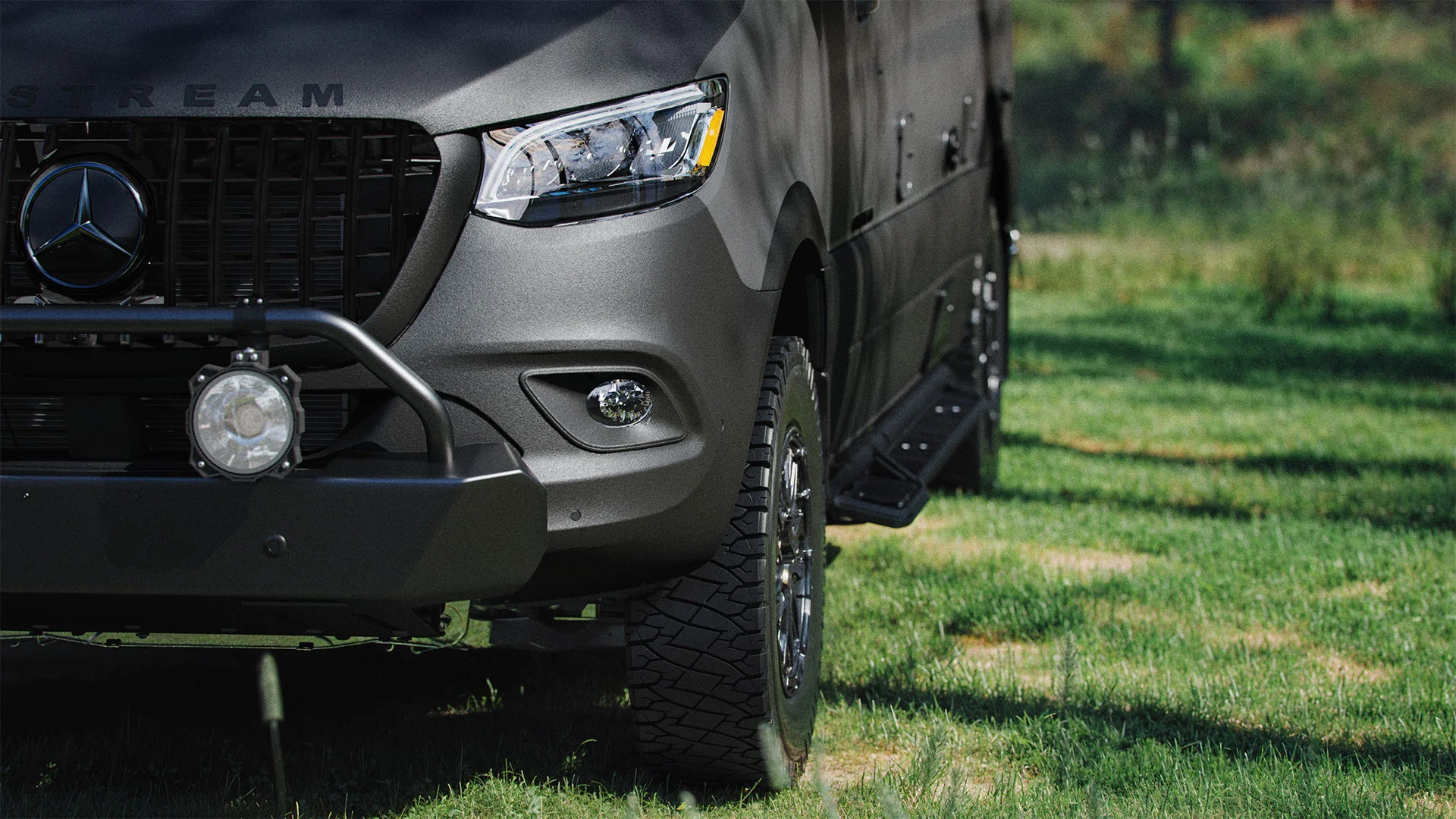 Close-up of the front right side of a matte black Mercedes-Benz van parked on grass, showing the grille, headlight, rugged tire, front bumper, and off-road accessory lights.