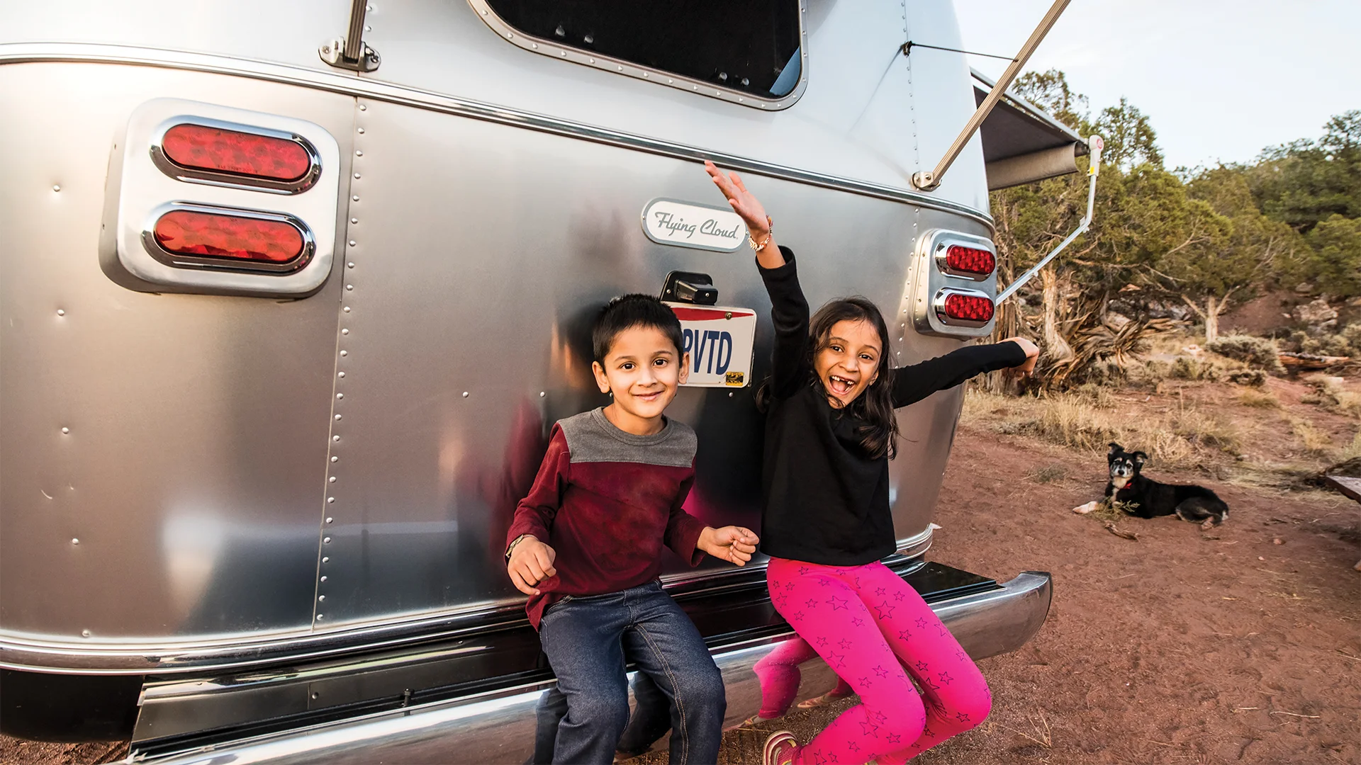 Two cheerful children sit on the bumper of a silver trailer with raised arms, while a dog rests in the background on a dirt ground.