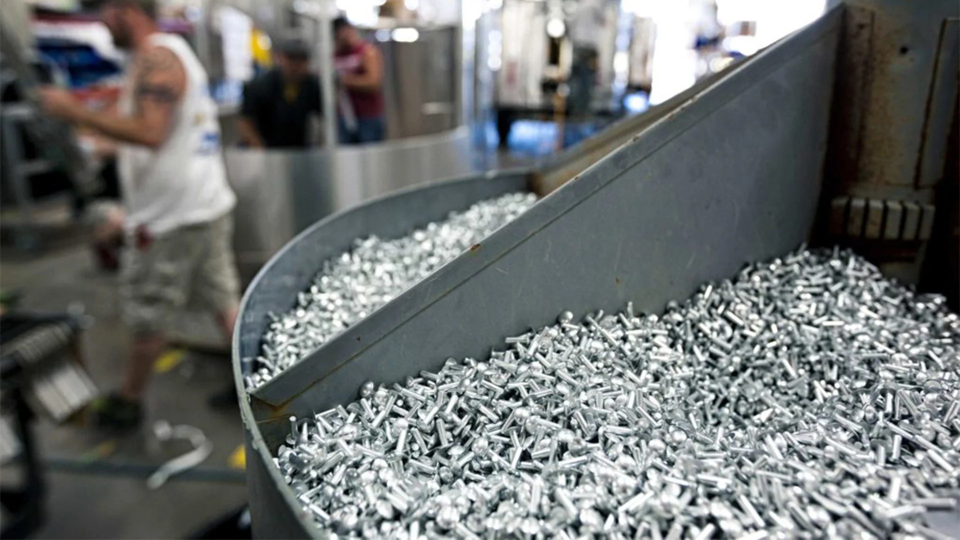 Close-up of a machine processing metallic pieces in a factory setting, with a worker in the background wearing a sleeveless shirt and shorts.