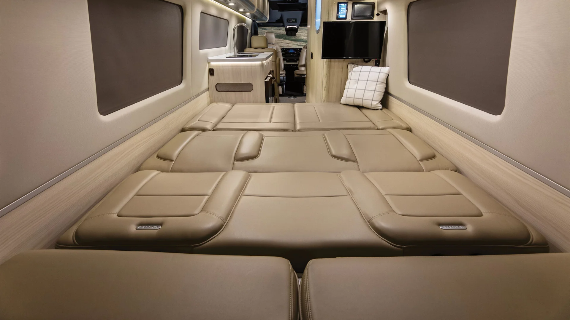 Interior of a luxury camper van with a fully extended bed in beige leather, recessed lighting, windows, and a wall-mounted screen.