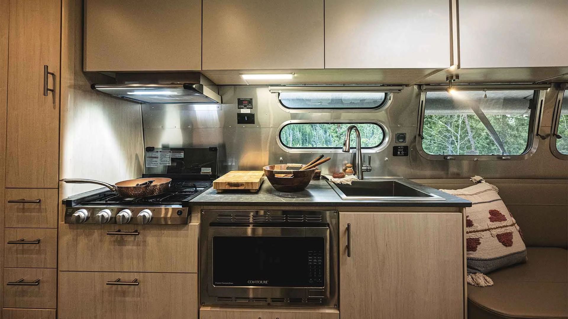 Modern RV kitchen with wood cabinets, stainless steel appliances, stove, microwave, sink, and window with nature view. Cozy pillows on sofa.