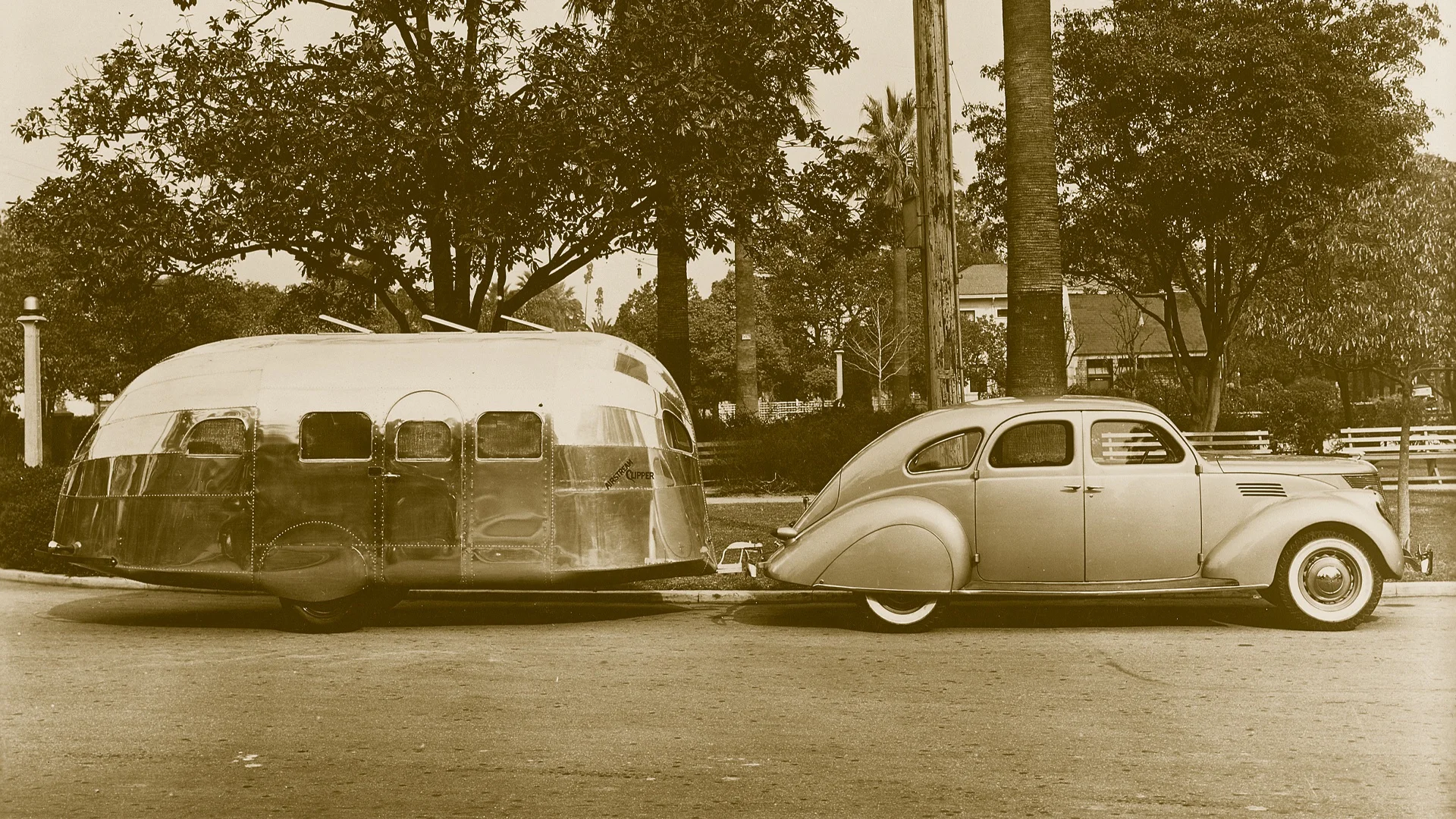 A vintage sepia-toned photo shows a streamlined 1930s car towing a shiny, rounded travel trailer, parked on a shaded street lined with trees and foliage.