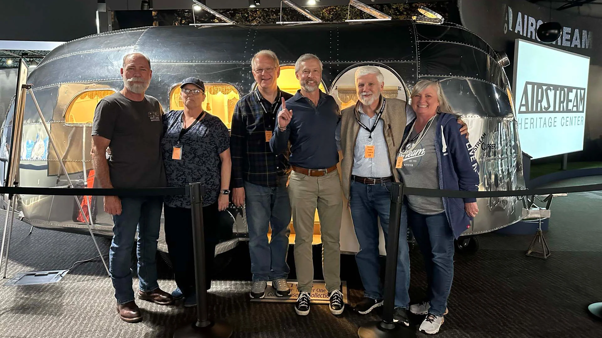 Group of six adults standing and smiling in front of a vintage Airstream trailer at a heritage center.