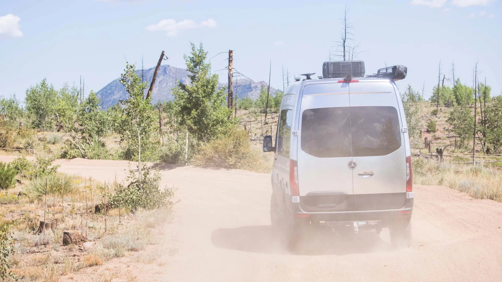 A silver van drives on a dusty dirt road surrounded by sparse trees, with a distant mountain under a clear blue sky.