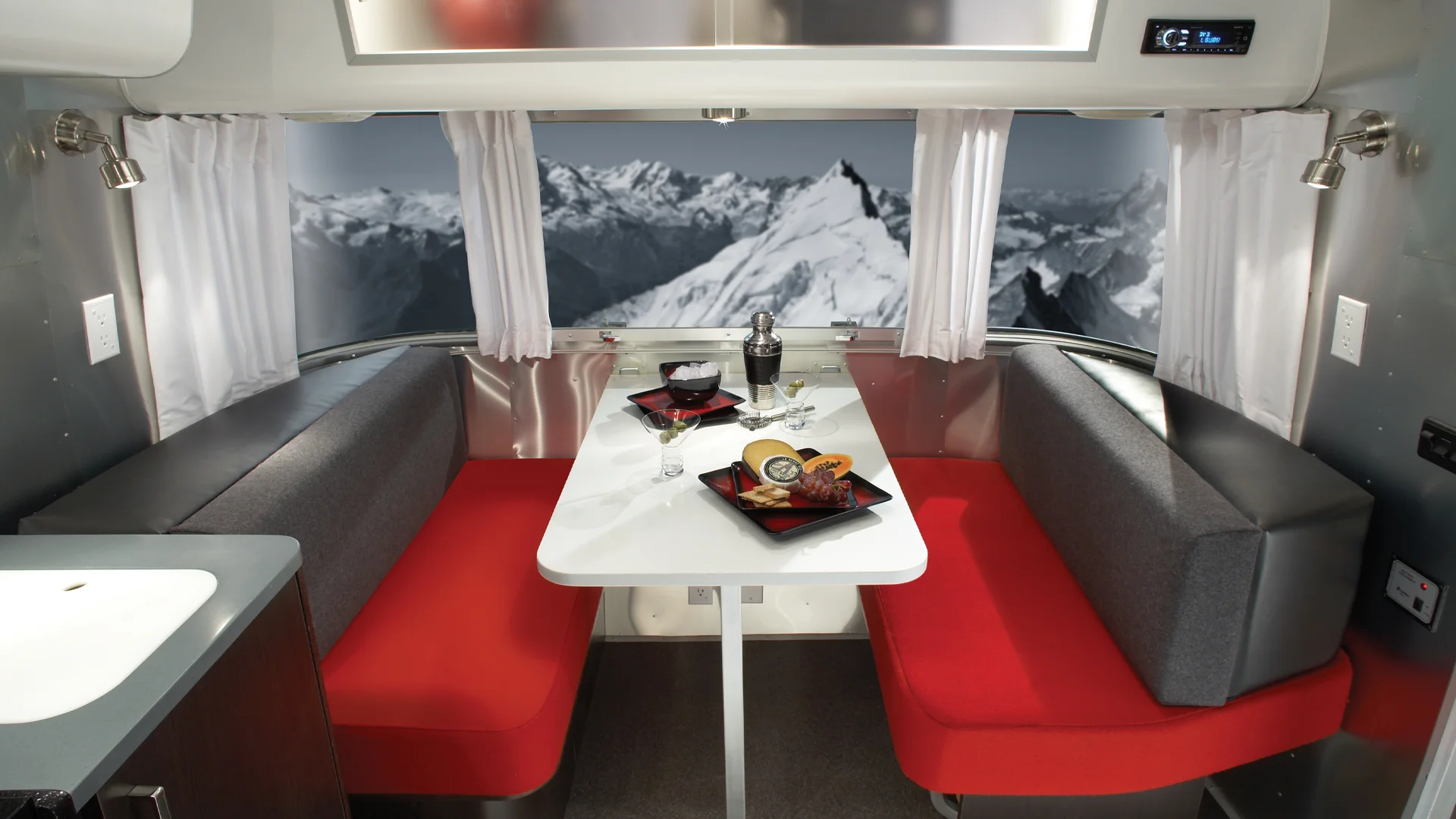 A modern RV dining area with red cushioned seats, a white table set with gourmet food, wine glasses, and a bottle, with large windows showing a scenic mountain landscape with snow-covered peaks.