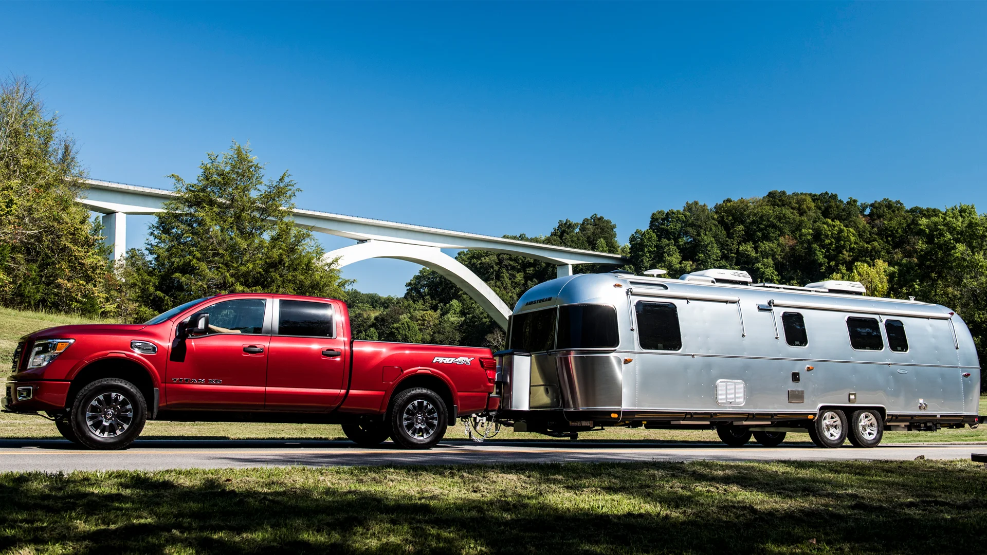 A red pickup truck is towing a silver Airstream travel trailer on a road, with green trees and a modern white bridge in the background under a clear blue sky.
