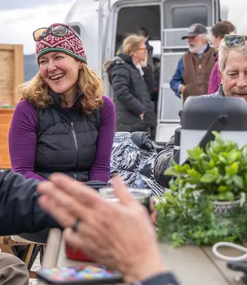 A group of people sit around an outdoor table, smiling and chatting. There are drinks, food, and a potted plant on the table. An Airstream trailer and other people are visible in the background.