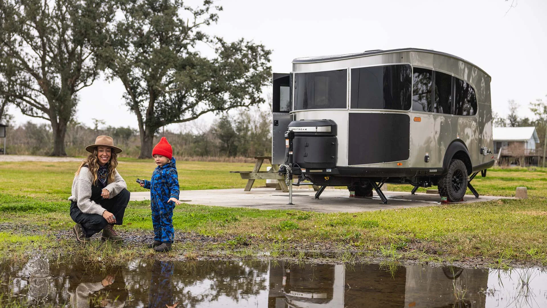 Woman and child in front of a modern travel trailer parked on grass. Trees and a small building in the background. The child wears a red hat.