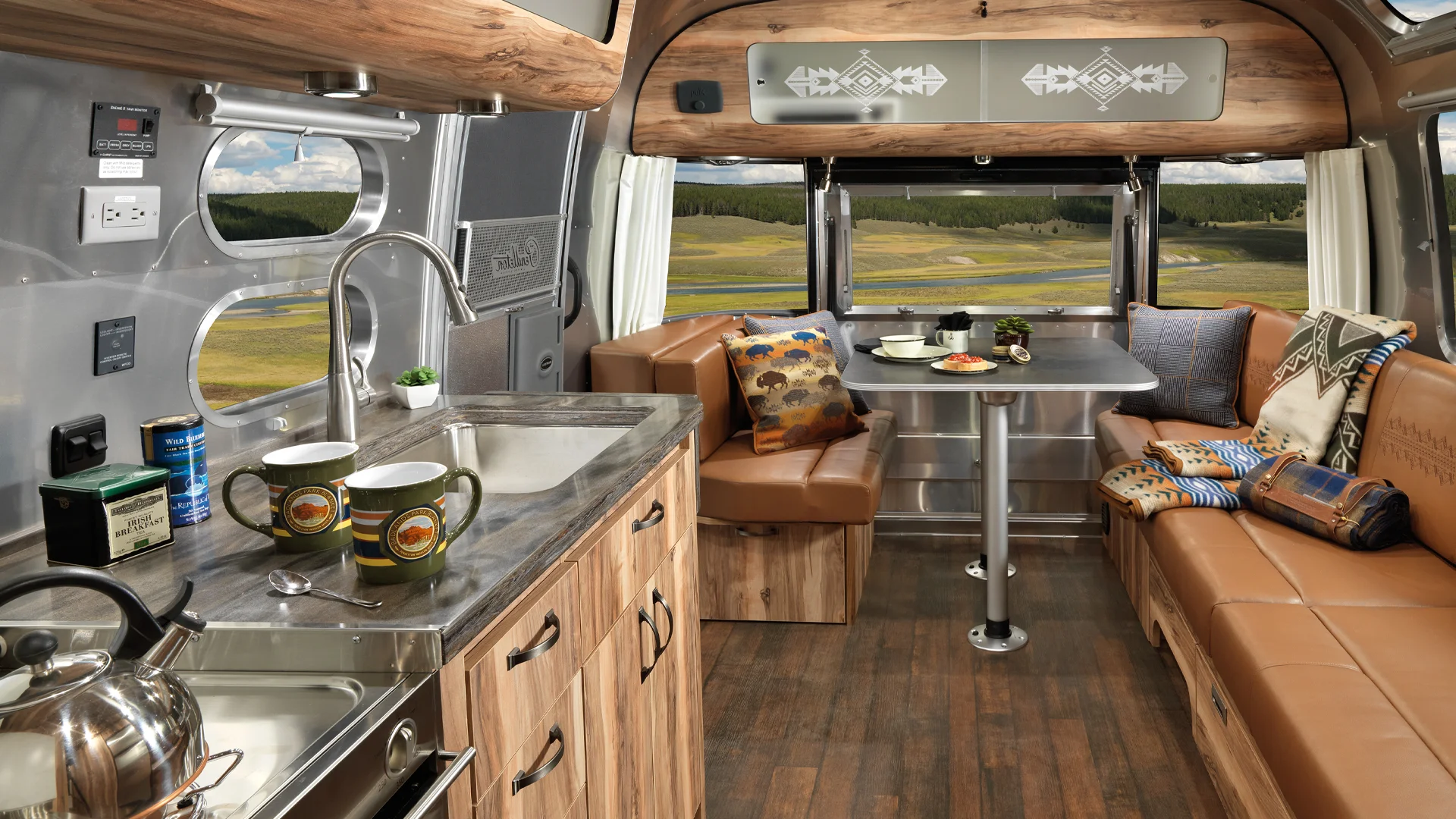 Interior of a modern camper van with wooden cabinets, leather seating, a table, and a view of scenic fields through large windows.