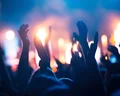 Crowd of people with raised hands enjoying a concert, with colorful stage lights illuminating the background in blue and orange hues.