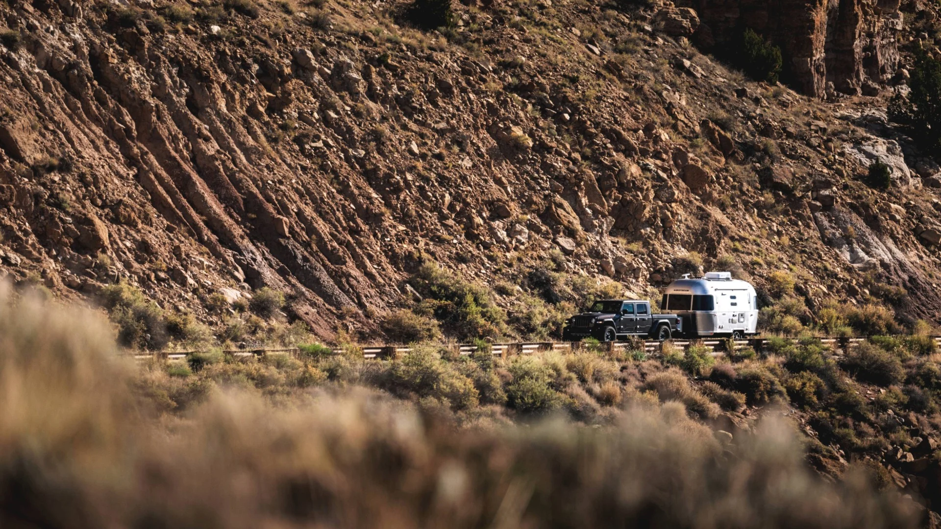 Truck towing a silver trailer along a rugged mountain road, surrounded by rocky hills and sparse vegetation.