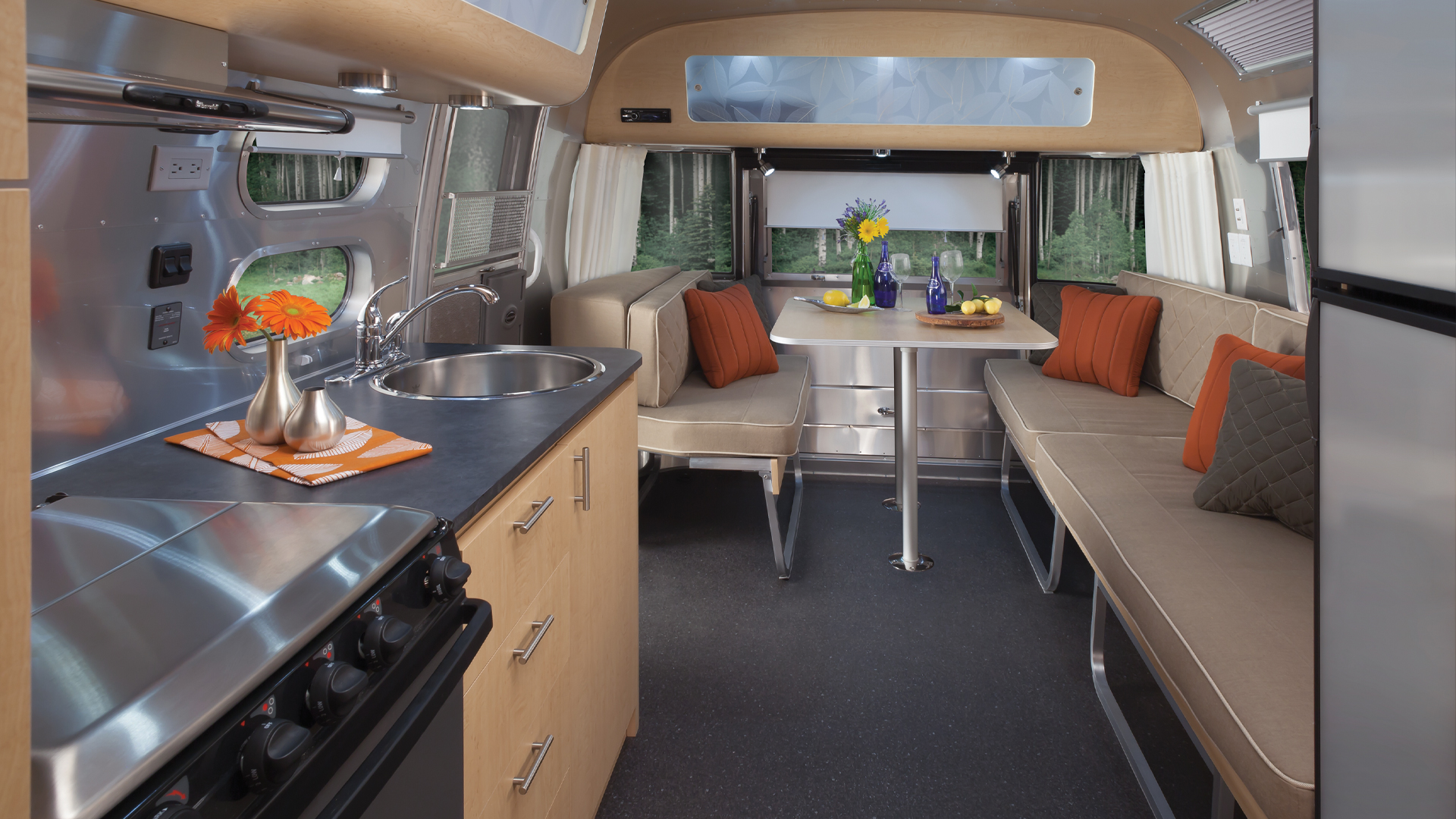 Interior of a modern camper van with a kitchen, dinette, and beige seating. A vase with flowers is on the table, and a forest view is outside.