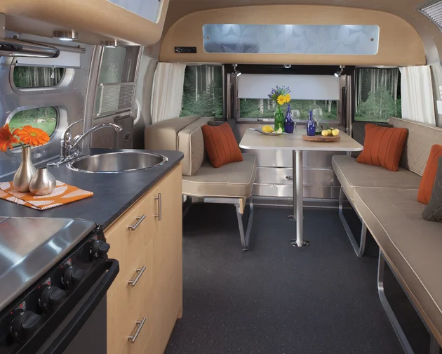 Interior of a modern camper van with a kitchen, dinette, and beige seating. A vase with flowers is on the table, and a forest view is outside.