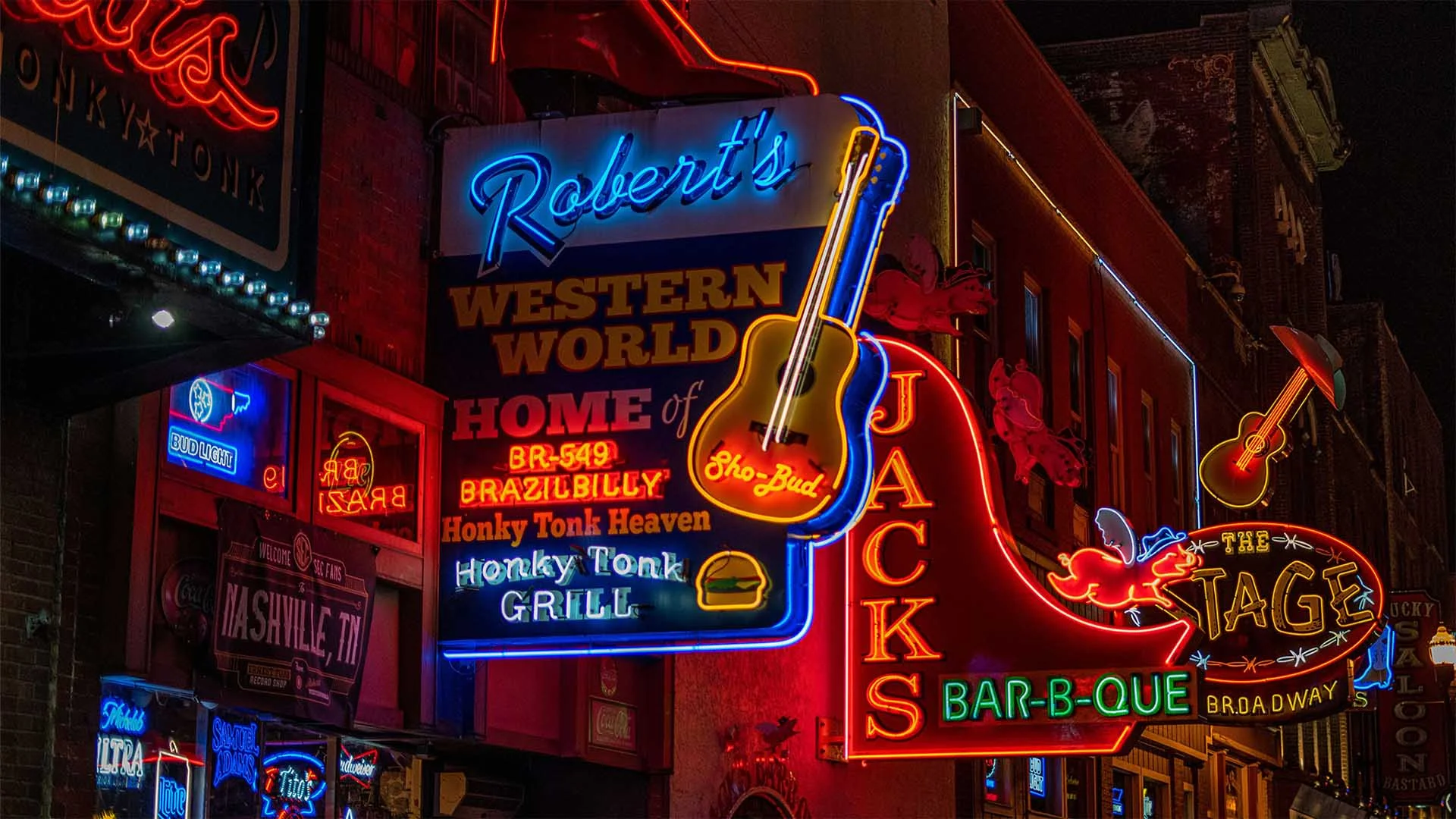 Neon signs for Robert's Western World, Jack's Bar-B-Que, and The Stage, featuring guitars and musical themes on a city street.