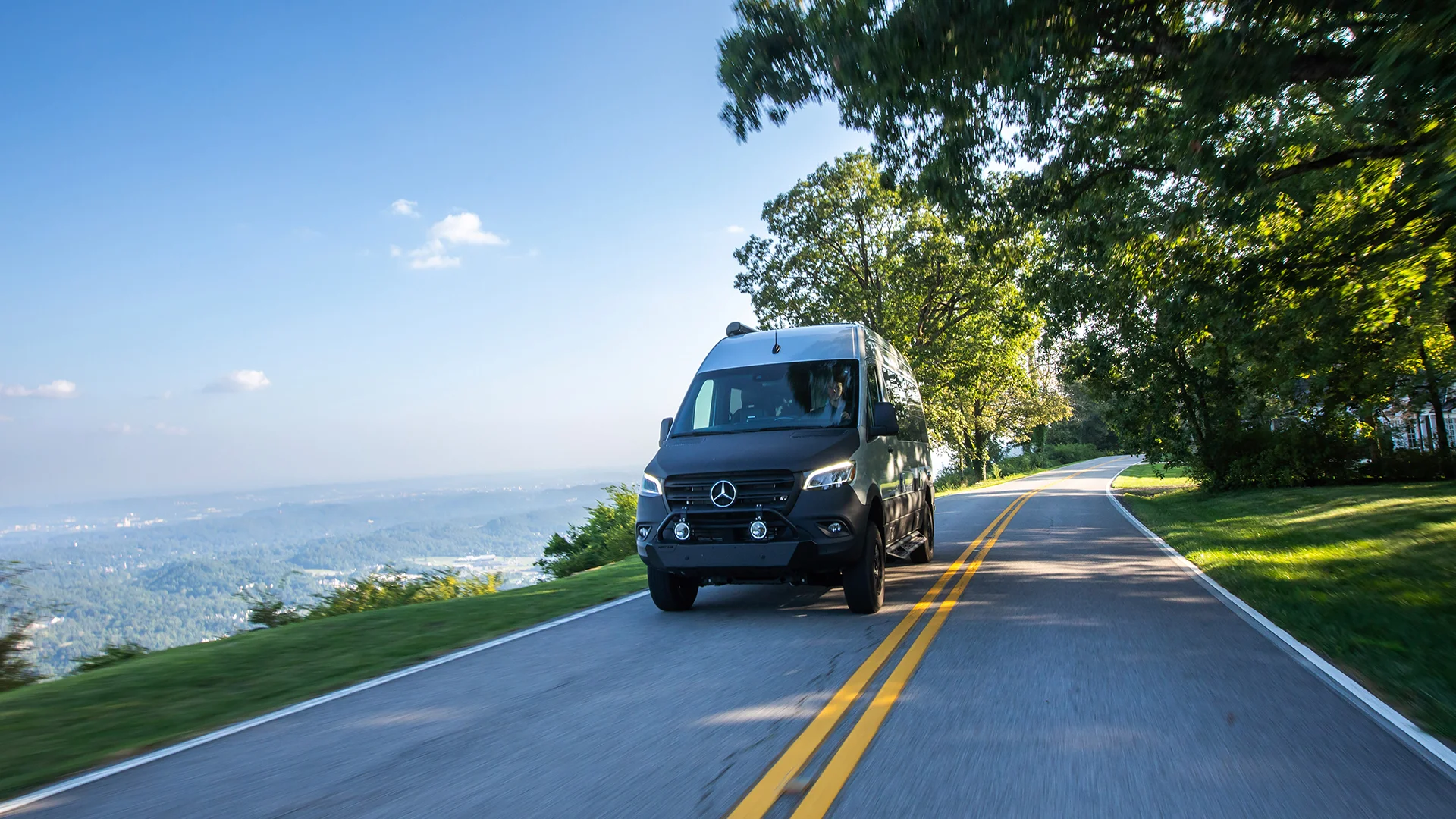 A black van drives on a scenic road flanked by trees, with a distant view of a valley under a clear blue sky.