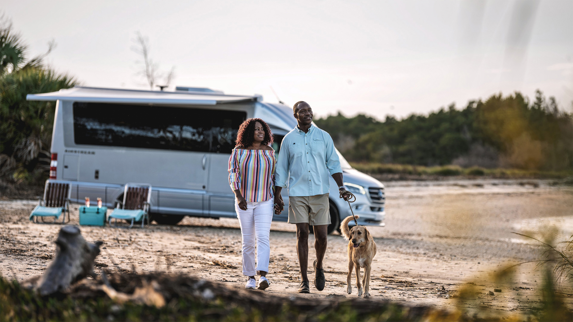 Couple walking a dog on a beach near a parked camper van, with trees in the background.