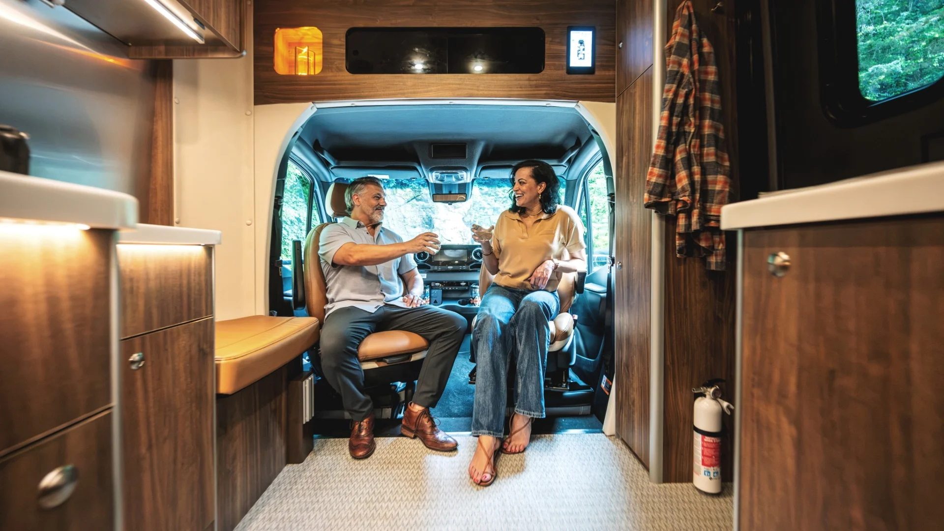 A man and woman sit in an RV interior, smiling and clinking glasses. The space is cozy with wood paneling and a window view of trees.