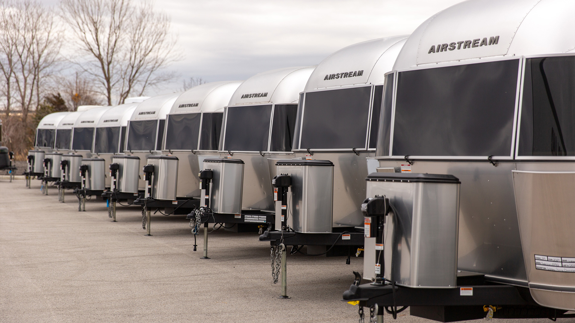 Row of silver Airstream trailers parked on concrete with bare trees in the background under a cloudy sky.