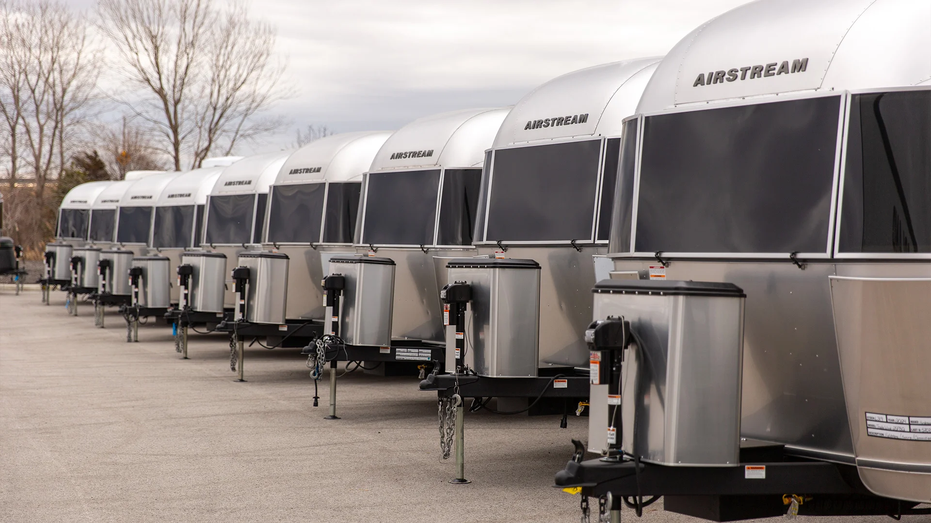 Row of silver Airstream trailers parked on concrete with bare trees in the background under a cloudy sky.