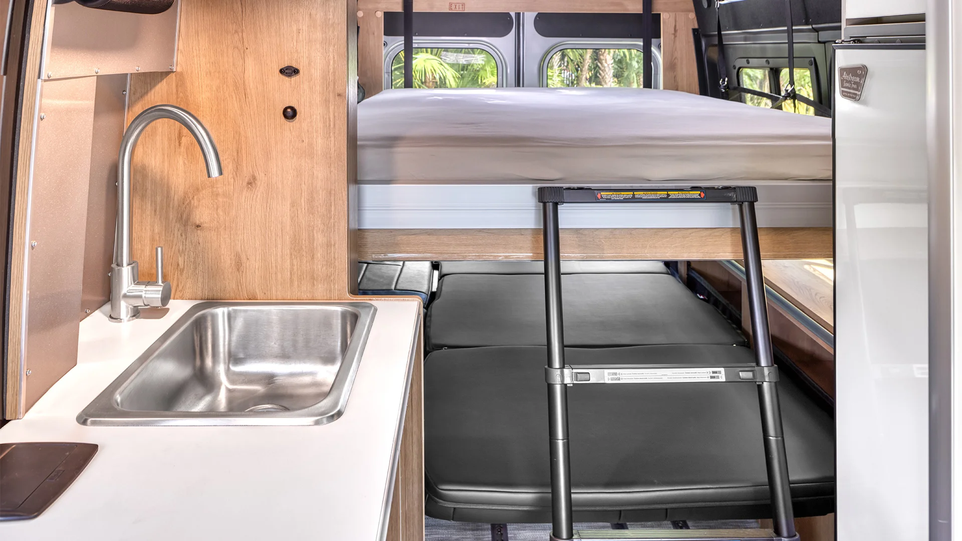 Compact camper van interior with a stainless steel sink, wooden accents, a ladder leading to an overhead bed, and a fold-out lower bed.