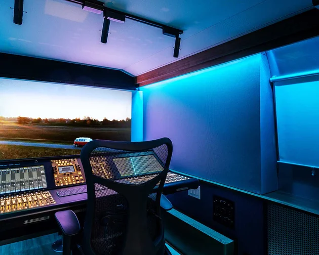 A modern sound mixing studio with a large control panel, mesh chair, and broad screen displaying a sunset landscape, lit by blue ambient lighting.