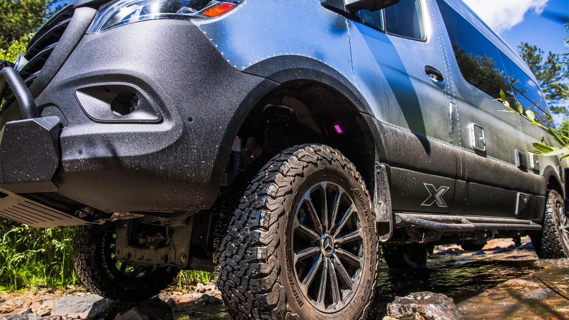 Close-up of an off-road vehicle with large tires, navigating a rocky terrain near a stream, surrounded by greenery and sunlight.