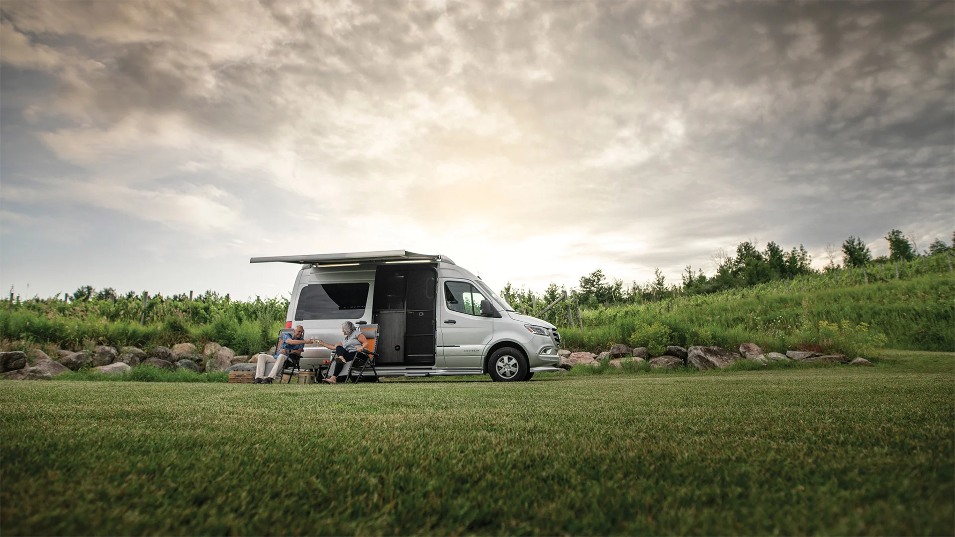 A camper van parked on a grassy field with two people sitting outside under an awning, enjoying the overcast skies and lush surroundings.