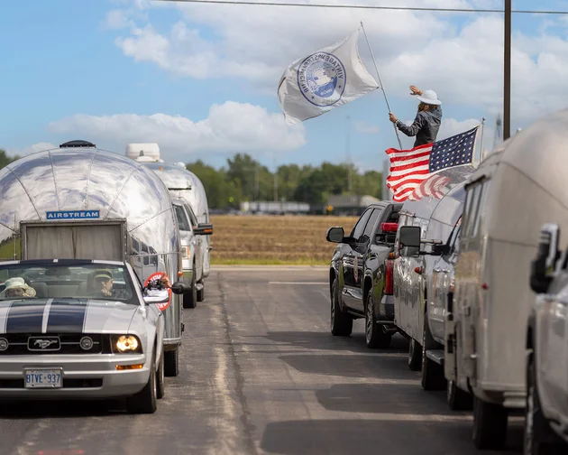 A convoy of Airstream trailers led by a Mustang car, with a person waving a flag from a vehicle, drives along a rural road.