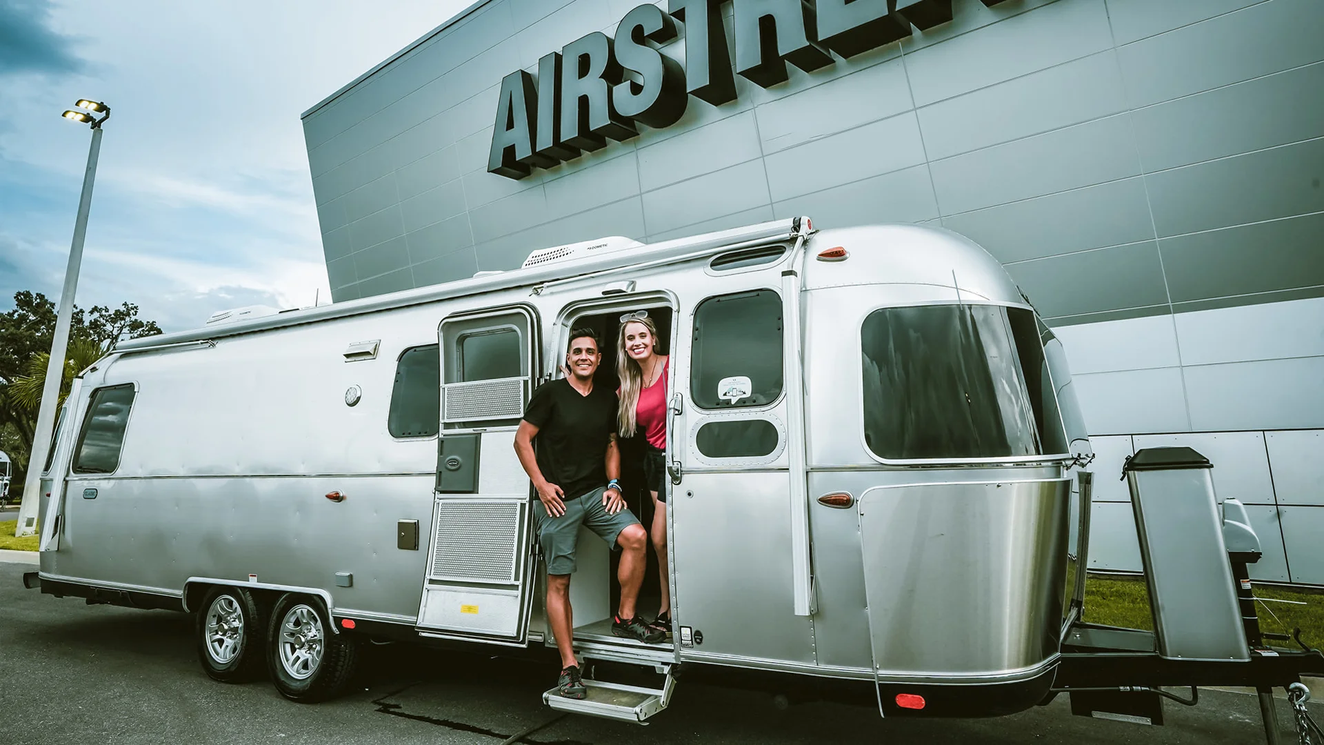 Two people stand smiling in the doorway of a shiny silver Airstream trailer parked in front of a modern building with the large word “AIRSTREAM” on it. The sky is partly cloudy.