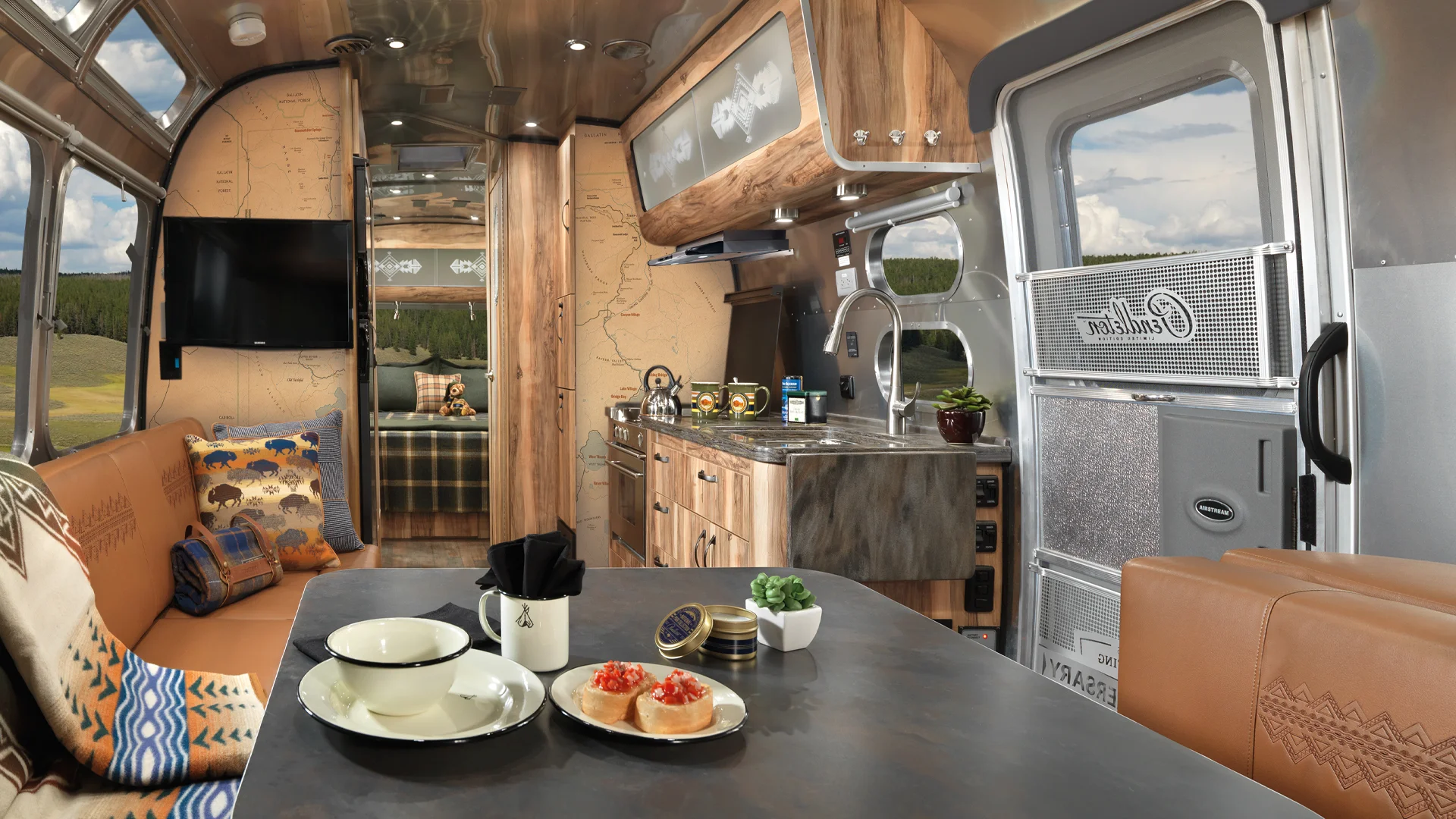 Modern Airstream trailer interior with wood accents, a seating area with patterned pillows, a dining table set with dishes and food, a compact kitchen, TV, and a bed in the background; outside is a scenic landscape.