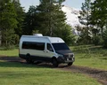 A white camper van drives along a gravel path amid lush trees, with snow-capped mountains visible in the background.