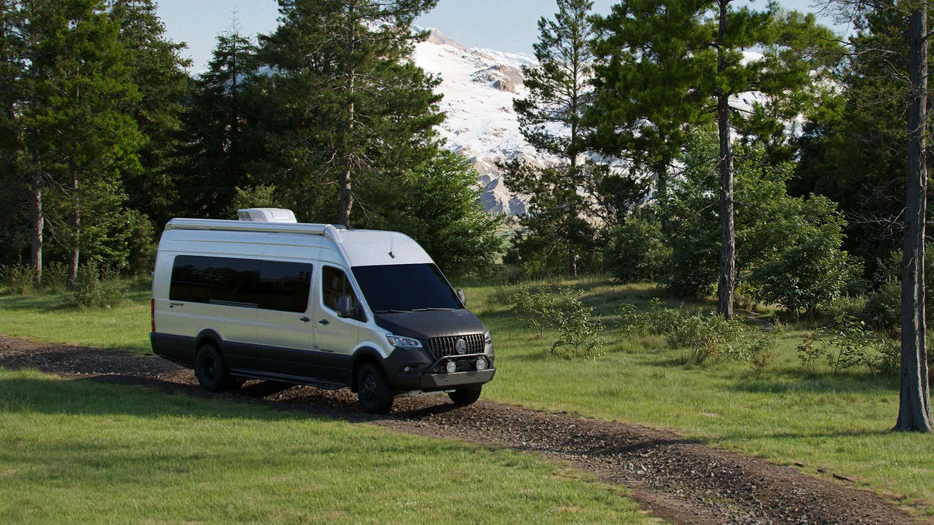 A white camper van drives along a gravel path amid lush trees, with snow-capped mountains visible in the background.
