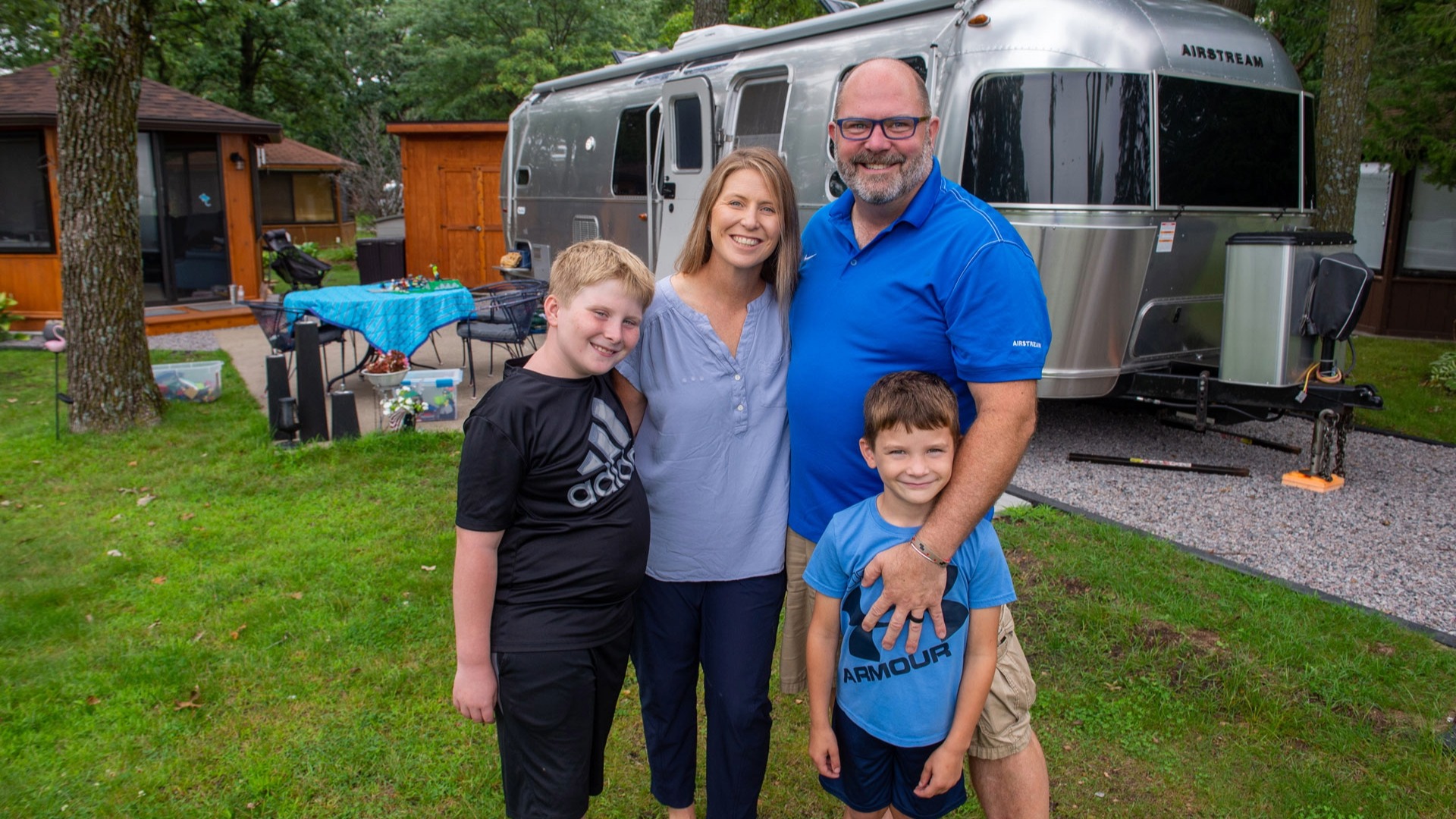 A family of four smiles outdoors, standing on grass beside a parked Airstream trailer and surrounded by trees and wooden cabins.