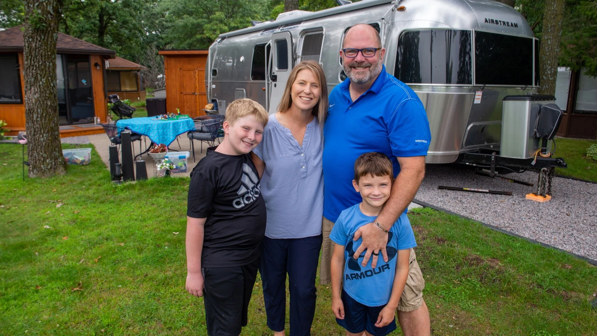 A family of four smiles outdoors, standing on grass beside a parked Airstream trailer and surrounded by trees and wooden cabins.