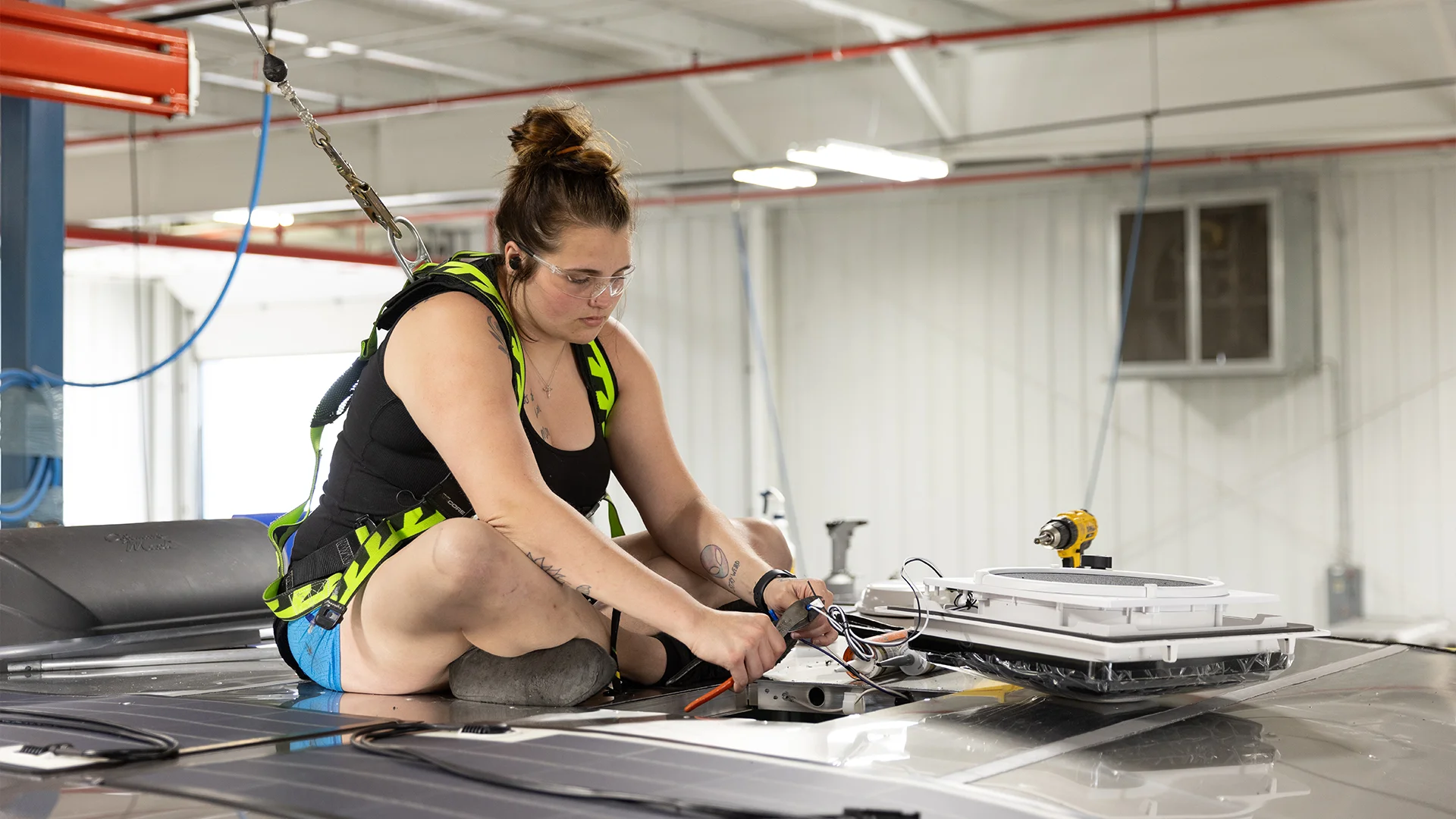 Person wearing safety gear installs equipment on a vehicle roof inside a bright, industrial workshop.