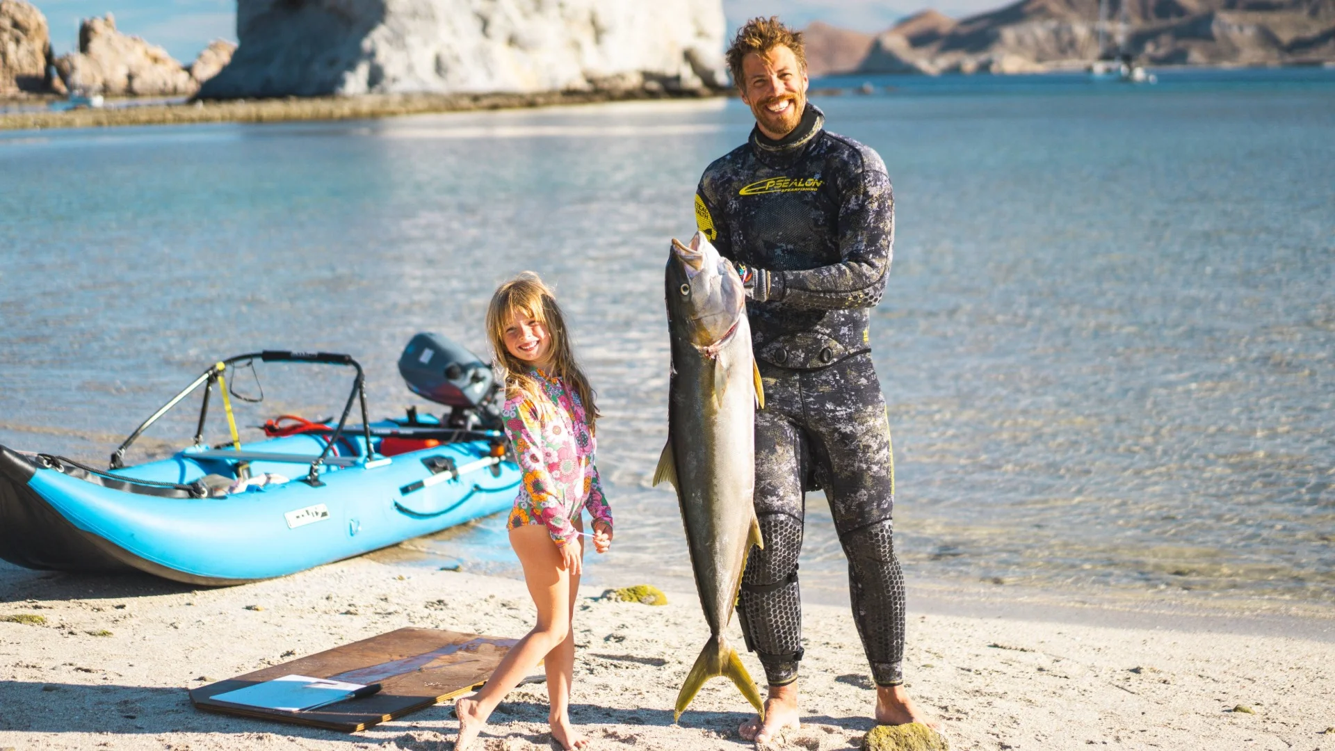 A man in a wetsuit and a child in a floral swimsuit stand on a beach holding a large fish, with a kayak beside them and the sea in the background.