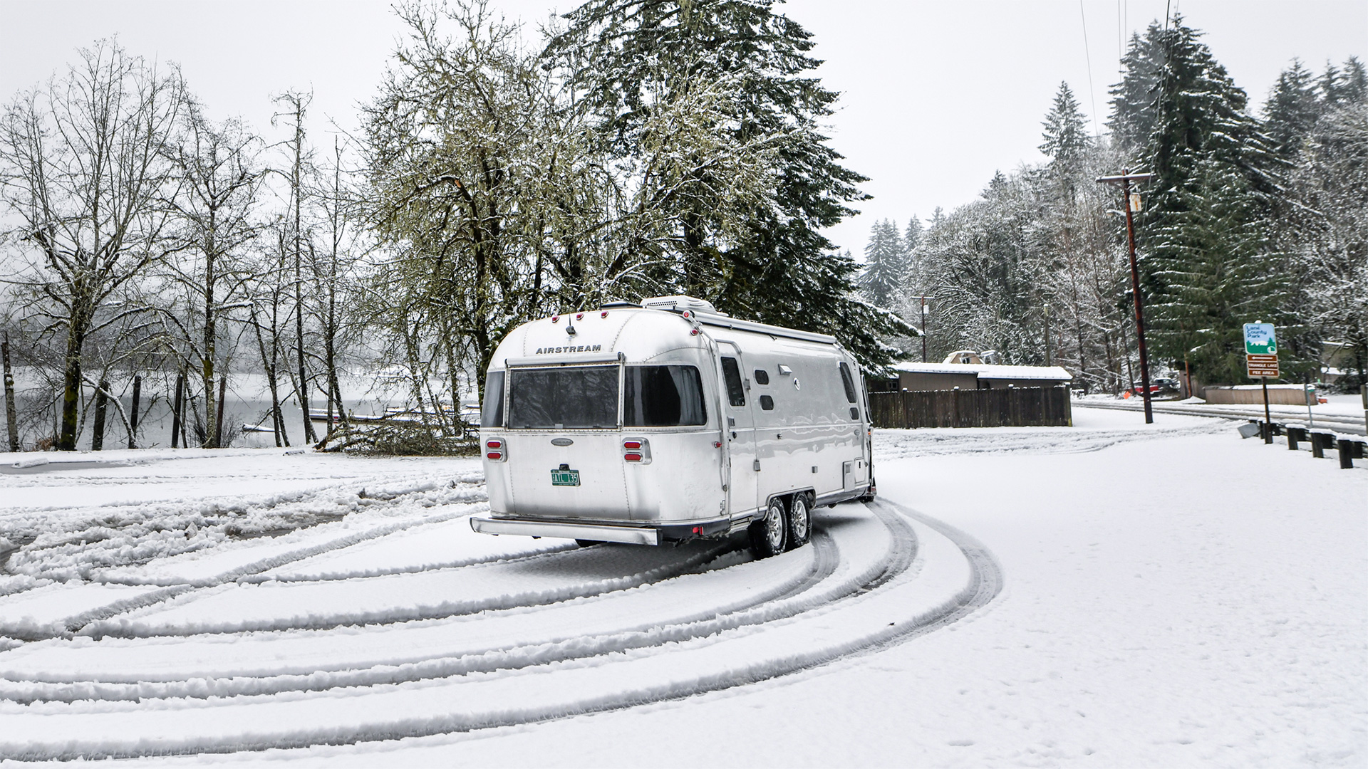 A silver Airstream trailer leaves tire tracks in fresh snow, surrounded by snowy trees and a lake in the background.
