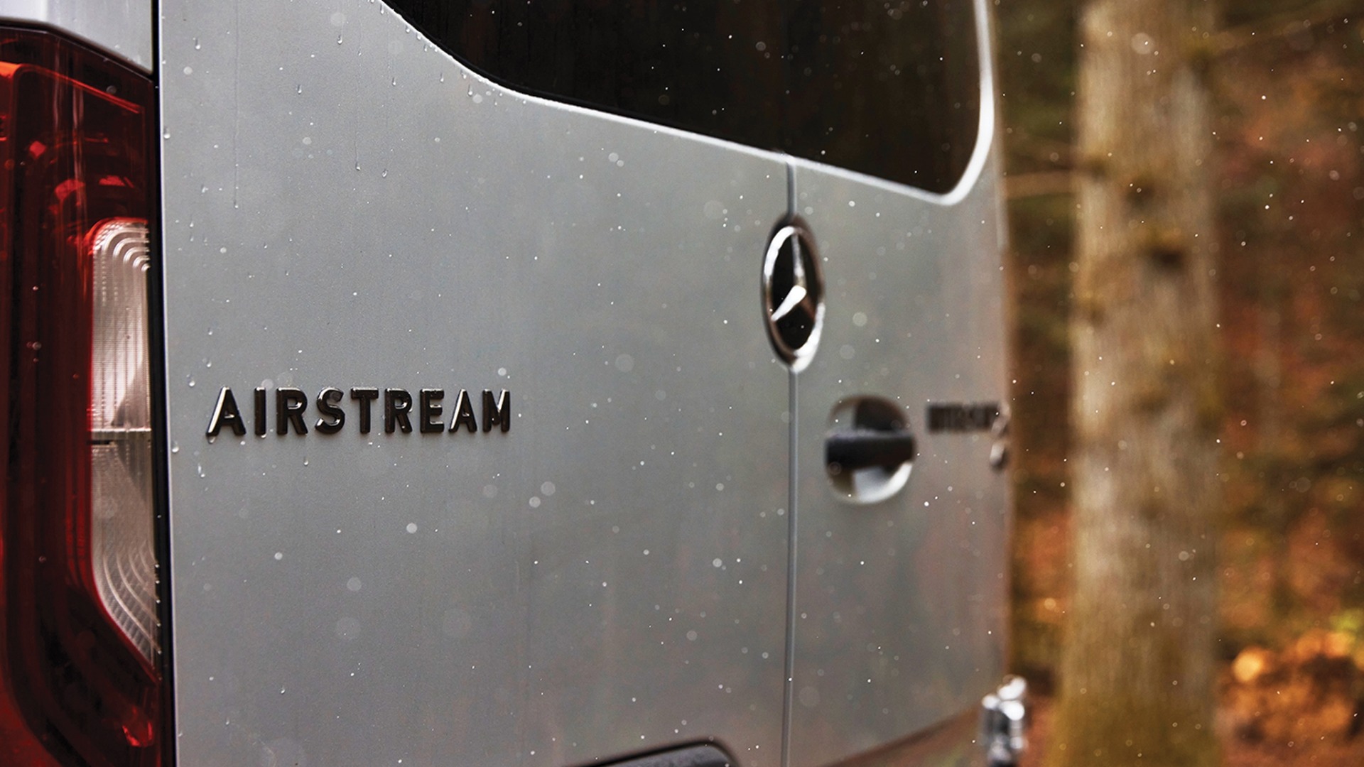 Close-up of a silver Airstream van's back, featuring a logo and taillight, with blurred trees in the background.