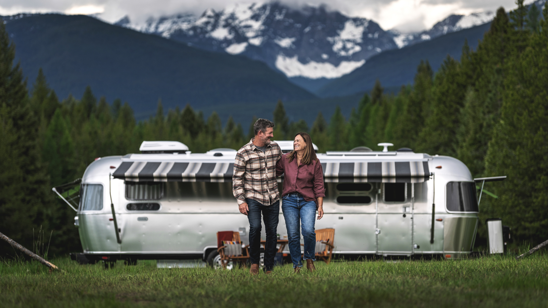 A couple walks hand in hand in front of a silver Airstream trailer, set against a backdrop of forested mountains under a cloudy sky.