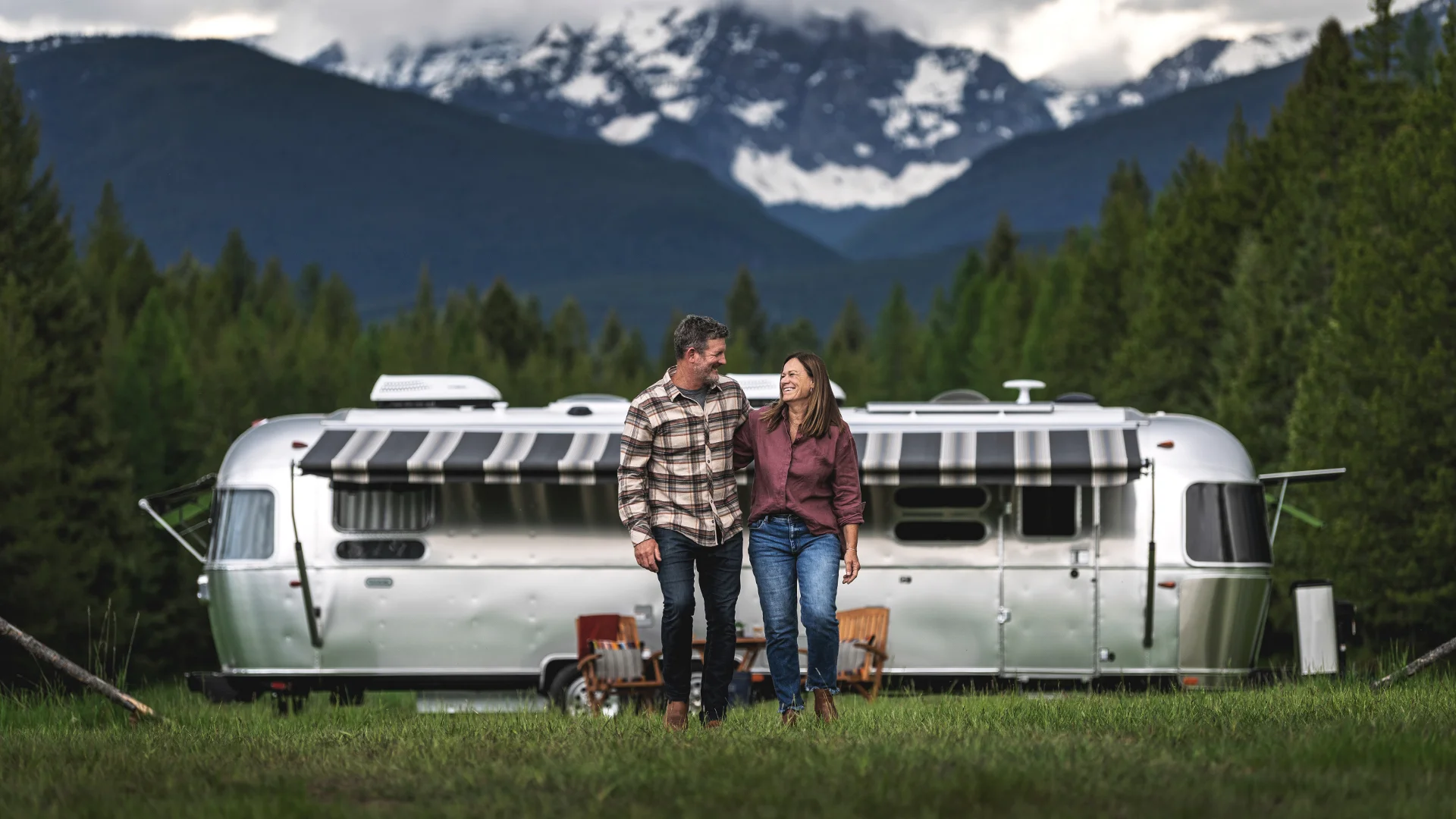 A couple walks hand in hand in front of a silver Airstream trailer, set against a backdrop of forested mountains under a cloudy sky.