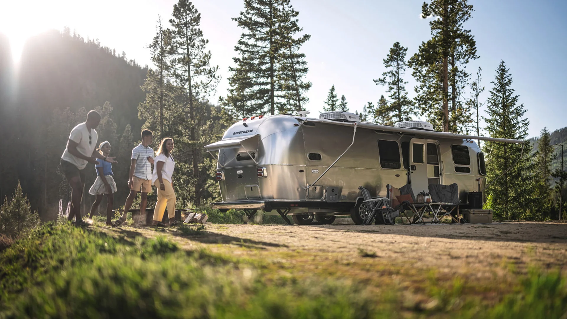 A family walks near a silver Airstream trailer in a forested area with mountains in the background, enjoying a sunny day.