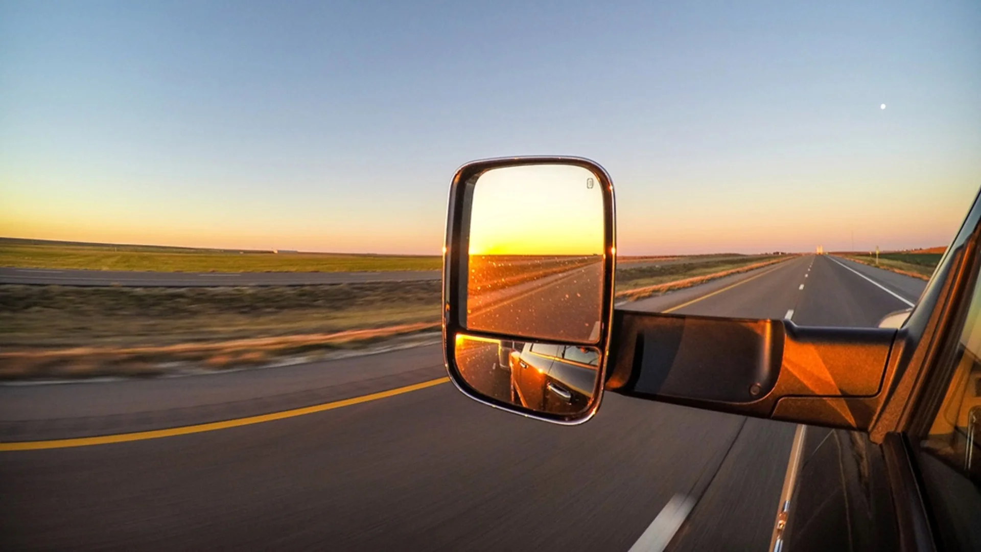 Side mirror view of a car driving on an open highway at sunset, reflecting the road and vivid orange sky.
