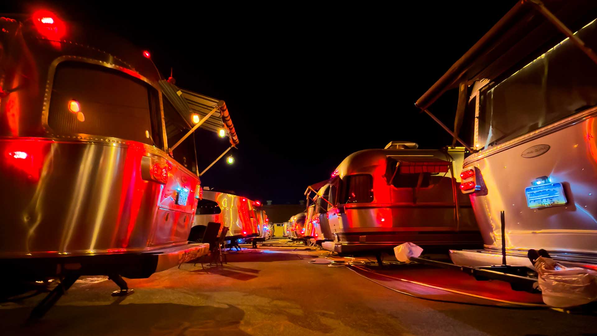 A nighttime scene of a line of brightly lit Airstream trailers parked closely together, illuminated by red and orange lights.