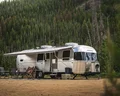 Silver Airstream trailer with an awning set up in a forested campsite, surrounded by chairs and a small table, with tall pine trees in the background.
