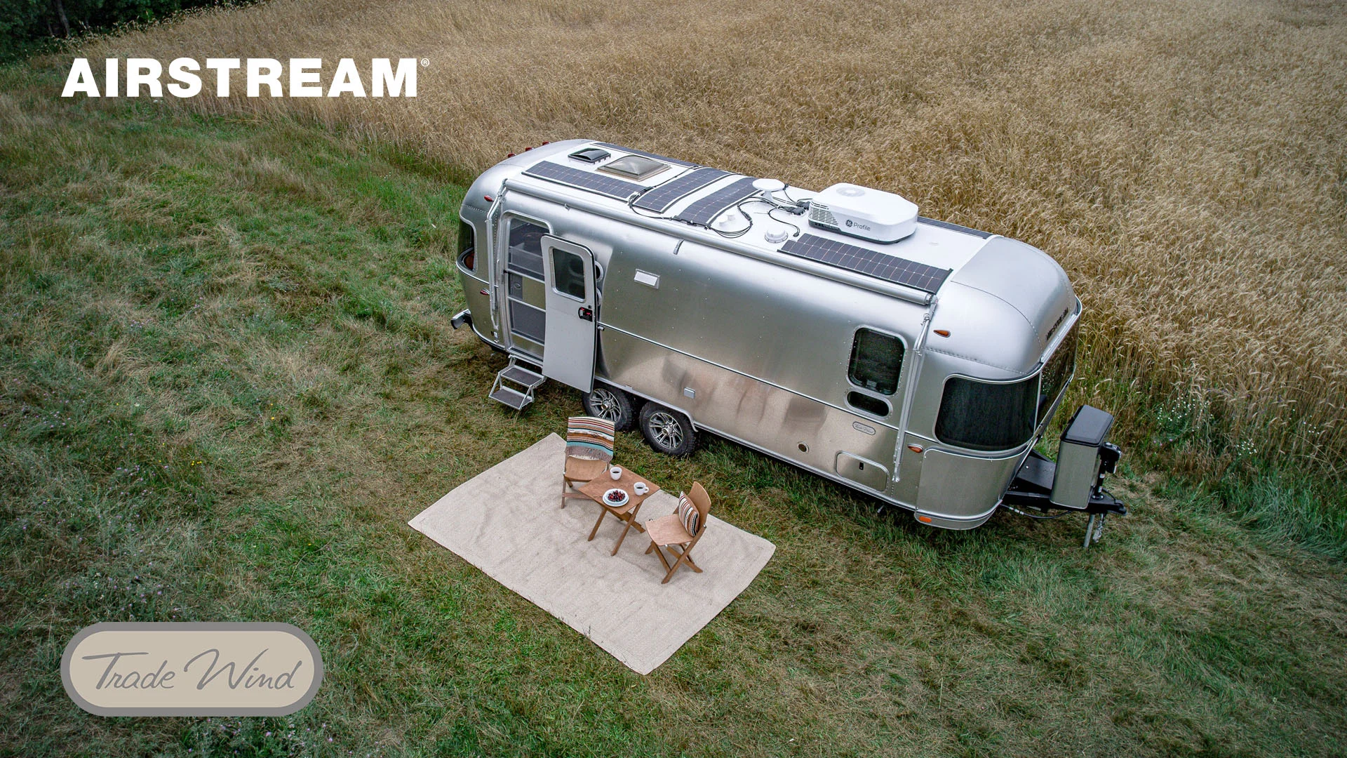Silver Airstream trailer in a grassy field with two chairs and a small table on a rug outside. Brand name "Airstream" visible above.