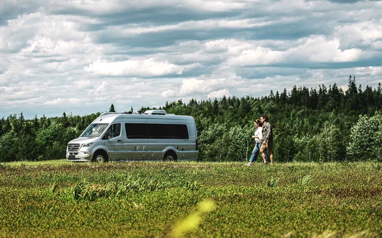 A couple walks hand in hand through a grassy field near a silver camper van, with a forest and cloudy sky in the background.