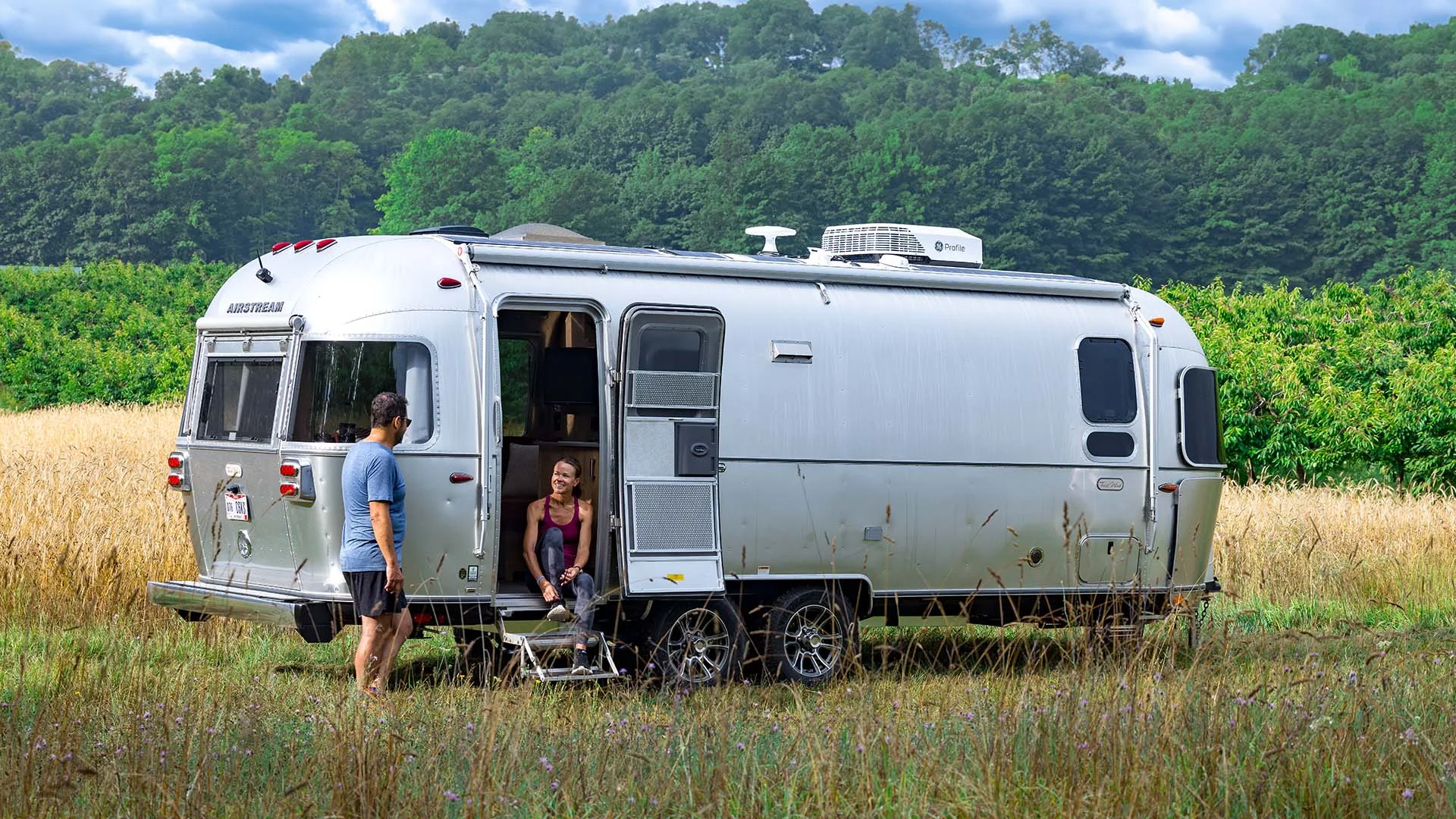 A shiny silver trailer in a grassy field with one person standing outside and another sitting inside, trees in the background.