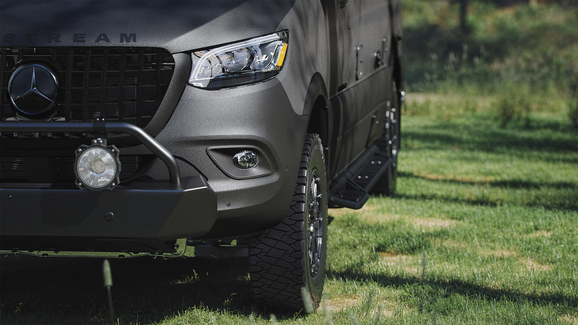 Front view of a matte black off-road vehicle with rugged tires parked on grass, showcasing a prominent grille and a mounted spotlight.