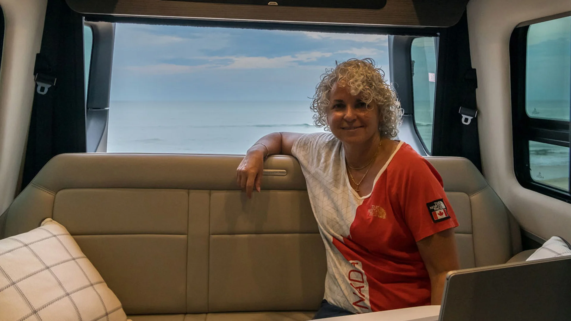 Person with curly hair sitting on a couch in a vehicle, with a window view of a cloudy sky and ocean.