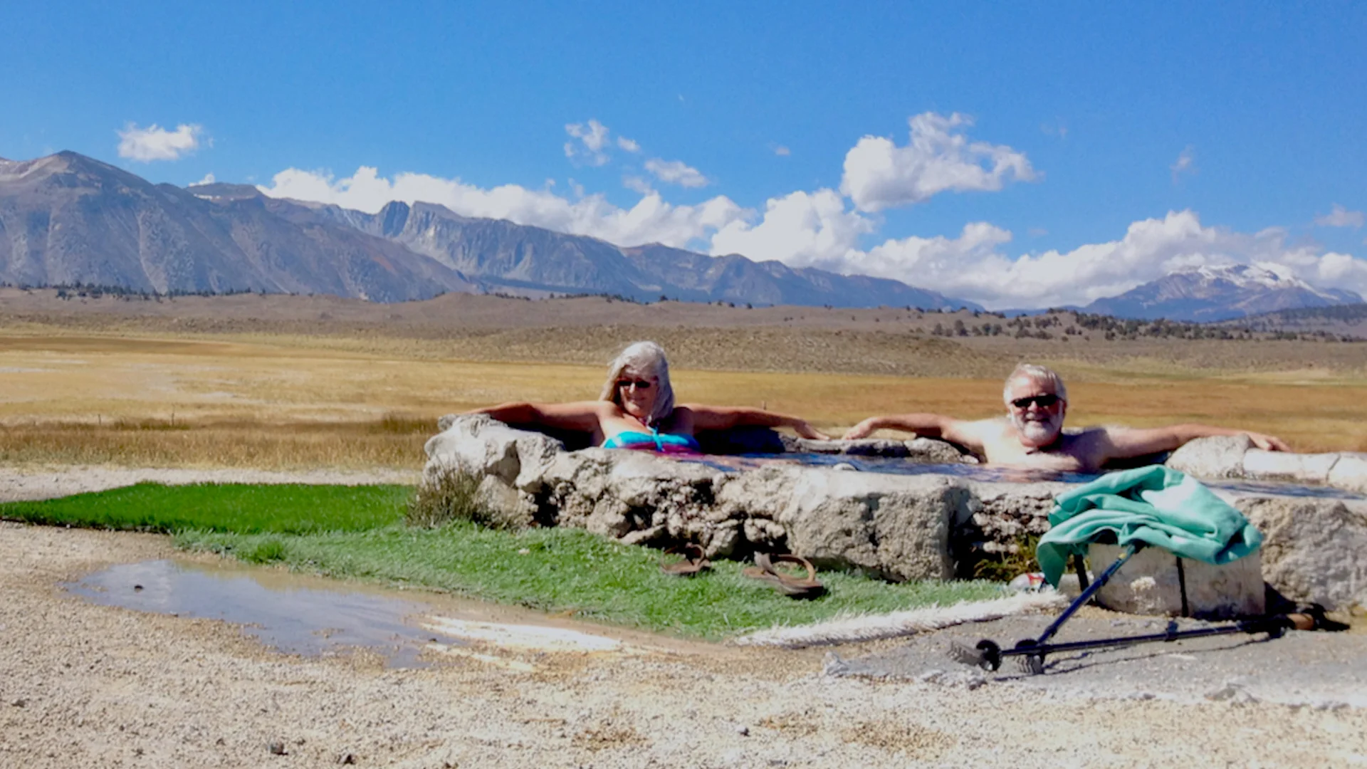 Two older adults relax in a stone hot spring set in an open, grassy plain with mountains in the background on a sunny day. A pair of walking poles and a backpack are resting nearby.